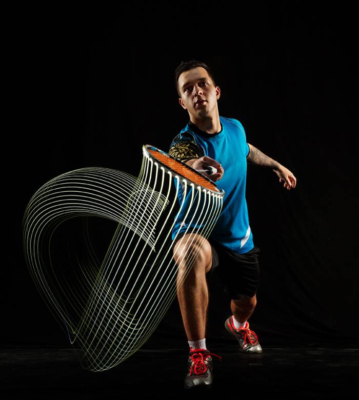 Young man playing badminton over black studio background. Fit male athlete isolated on dark with led light trail . badminton player in action, motion, movement. attack and defense concept